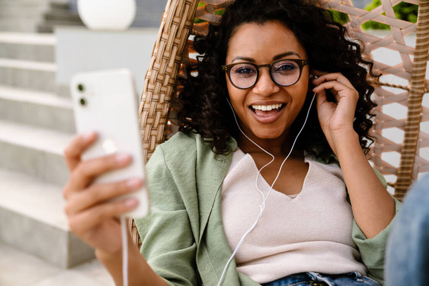 Excited smiling young african american woman in earphones using smartphone while sitting outdoors on rattan hang chair - Photo, Image