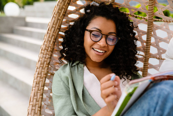 Young smiling african woman in eyeglasses writing down notes while sitting outdoors on rattan hang chair - Zdjęcie, obraz