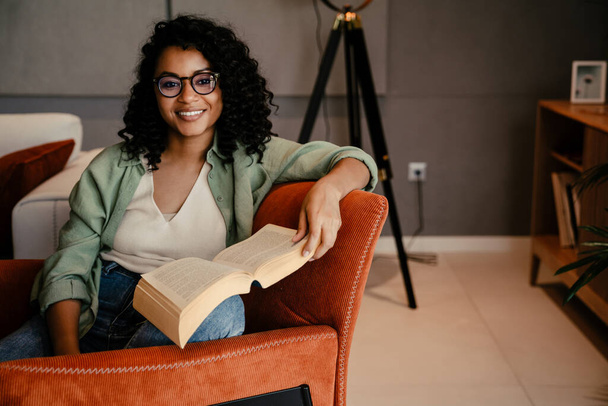 Young smiling african woman in eyeglasses reading book while sitting on armchair at home - Foto, Imagem