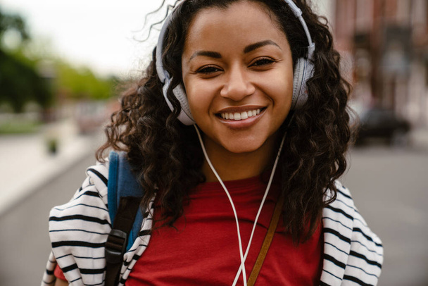 Young black woman in headphone looki camera while walking outdoors - Foto, afbeelding
