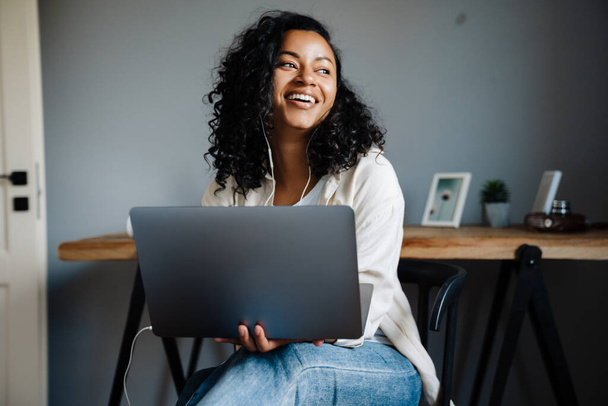 Black young woman smiling and working with laptop while sitting on chair at home - Zdjęcie, obraz