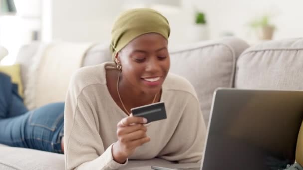 Kannettava tietokone, luottokortti ja innoissaan musta nainen sohvalla tehdessään verkko-ostoksia kotonaan. Happy, verkkokauppa ja Afrikkalainen nainen ostaa vaatteita vähittäiskaupan verkkosivuilla tietokoneella samalla rentouttava - Materiaali, video