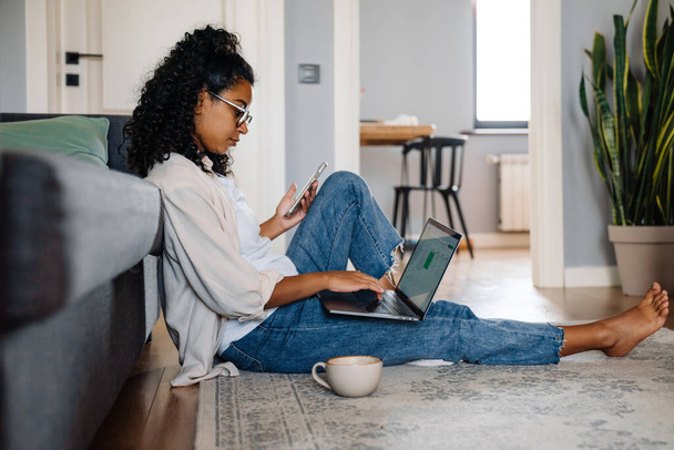 Black young woman using gadgets and drinking coffee while sitting on floor at home - Φωτογραφία, εικόνα