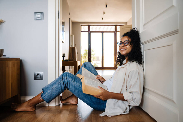 Black young woman in eyeglasses reading book while sitting on floor at home - Fotó, kép