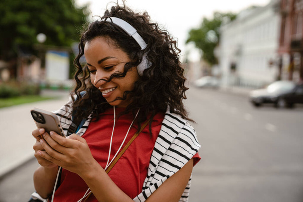 Young black woman in headphones using mobile phone while walking outdoors - Photo, image