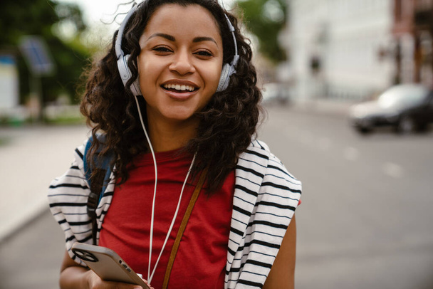 Young black woman in headphones using mobile phone while walking outdoors - Photo, Image