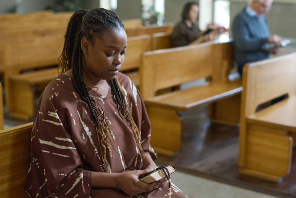 説教中に福音主義教会のベンチに座っている間、聖書とバラ色のビーズを持っている若いアフリカ系アメリカ人女性 - 写真・画像