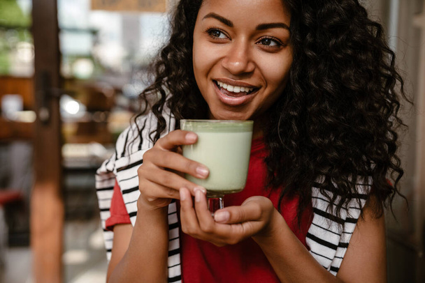 Young black woman smiling and drinking matcha latte while sitting at cafe outdoors - Фото, зображення