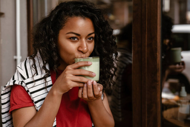 Young black woman drinking matcha latte while sitting at cafe outdoors - Fotoğraf, Görsel