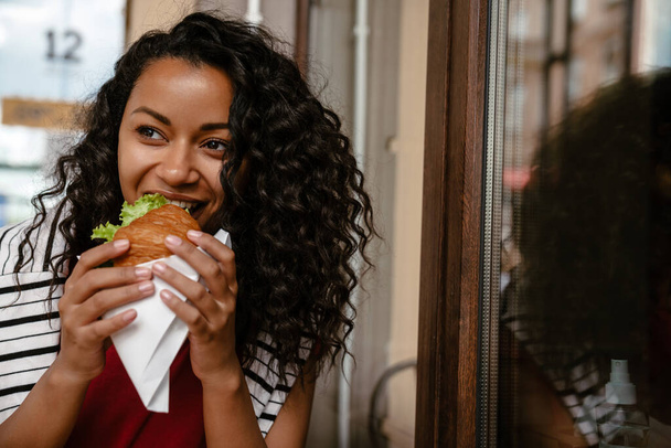 Young black woman smiling and eating sandwich while sitting at cafe outdoors - Foto, imagen