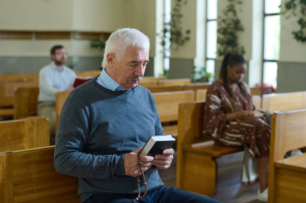 Vážné šedovlasý muž v casualwear držení Bible a růženec korálky před sebou při tiché modlitbě proti černošce v kostele - Fotografie, Obrázek