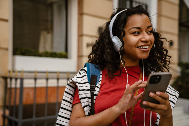 Young black woman in headphones using mobile phone while standing outdoors - 写真・画像