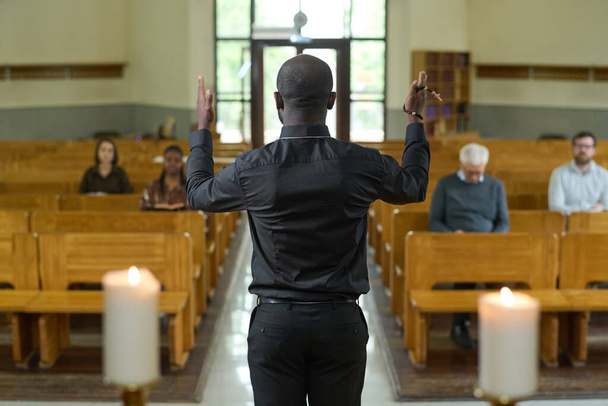 Visão traseira do jovem padre afro-americano em camisa preta e calças abençoando paroquianos sentados em bancos durante o serviço da igreja - Foto, Imagem