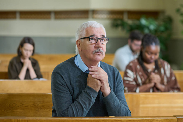 Alter Mann in Freizeitkleidung und Brille hält die Hände während des stillen Gebets zusammen, während er das Kreuz in der Kirche betrachtet - Foto, Bild