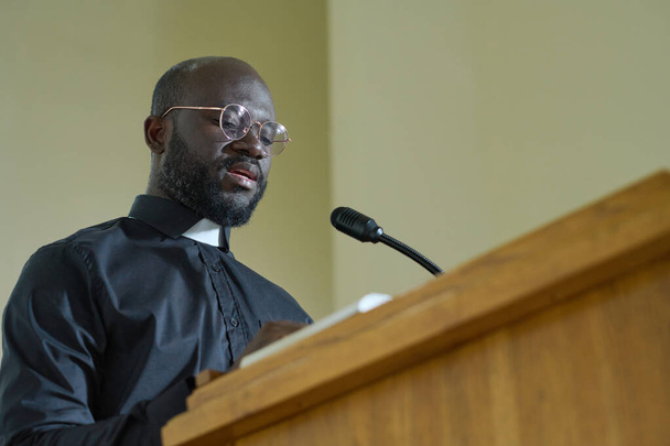 Young priest in black shirt with clerical collar reading verses from Bible and explaining them while pronouncing speech by pulpit - Fotó, kép