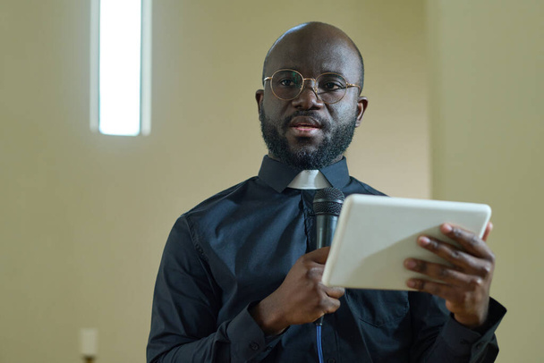 Nuori itsevarma musta mies, jolla on mikrofoni ja digitaalinen tabletti, joka pitää puheen evankelisen kirkon seurakuntalaisille saarnan aikana - Valokuva, kuva