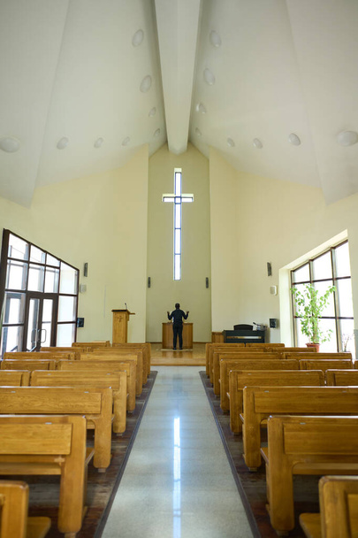 Langer Gang zwischen zwei Reihen hölzerner Bänke, die zum Priester führen, der vor dem Altar steht und mit erhobenen Armen auf das Kreuz blickt - Foto, Bild
