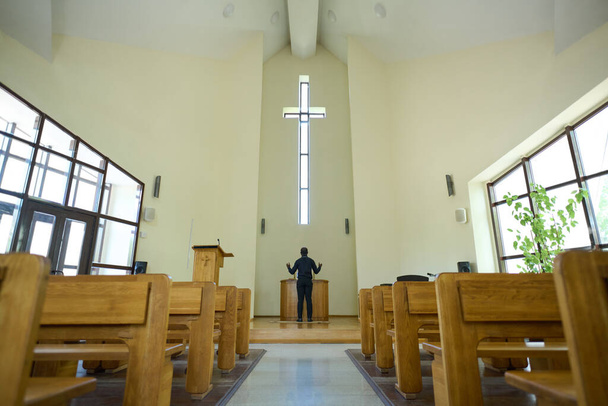 Interiér kostela s dlouhou uličkou vedoucí k pastorovi, který stojí před křížem a dřevěnou kazatelnou a drží ruce v požehnání - Fotografie, Obrázek