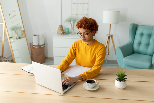Chica afroamericana que usa el ordenador portátil en la oficina en casa mirando la pantalla escribiendo chateando leyendo el correo electrónico de escritura. Mujer joven que tiene una reunión virtual de chat en línea de videoconferencia. Aprendizaje laboral desde casa - Foto, imagen