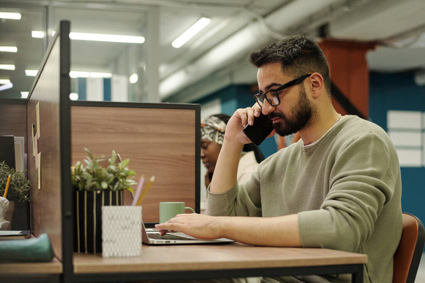 Fiatal, komoly szemüveg és alkalmi viselet dolgozó beszél az ügyféllel mobiltelefonon, miközben ül a munkahelyén, és nézi a laptop képernyőn - Fotó, kép