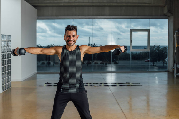 Portrait of a smiling caucasian male personal instructor, looking at camera, exercising with dumbbells in a big gym. workout lifting dumbbell weights. Horizontal, window background, Copy-space. - Photo, Image