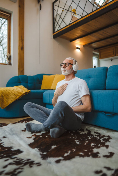 Ein Mann Senior kaukasischen männlichen Augen geschlossen für geleitetes Training Yoga oder Meditation, während zu Hause sitzen mit Kopfhörern Selbstpflege üben echte Menschen Wohlbefinden inneren Frieden und Gleichgewicht Konzept - Foto, Bild