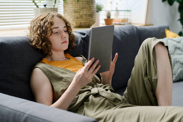 Молодая расслабленная белая женщина в повседневной одежде просматривает онлайн-данные на экране цифрового планшета, отдыхая дома на диване - Фото, изображение