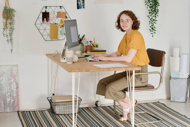 Onnellinen nuori nainen freelancer casualwear istuu pöydän ääressä tietokoneen monitorin edessä olohuoneessa ja katselee kameraa - Valokuva, kuva