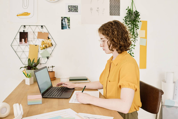 Молода серйозна бізнес-леді в повсякденному одязі сидить за столом перед ноутбуком, роблячи нотатки у фінансовому документі та аналізуючи дані
 - Фото, зображення