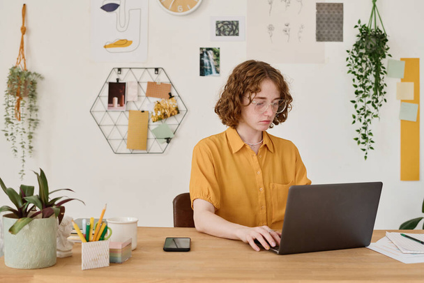 Молодий студент або фрілансер в повсякденному одязі сидить перед ноутбуком за робочим місцем вдома і дивиться онлайн-дані на екрані
 - Фото, зображення