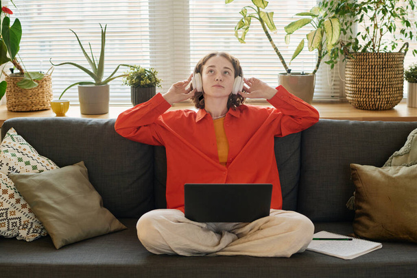 Young female student in headphones listening to audio lesson while sitting on couch in living room with laptop on her crossed legs - 写真・画像