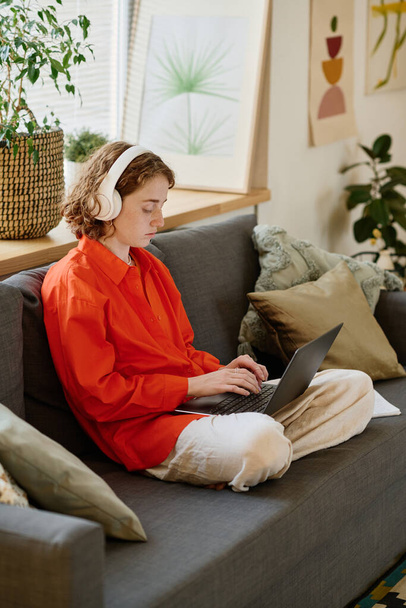 Fiatal lány diák piros ingben és fehér nadrág gépelés laptop billentyűzeten, miközben ül a puha kényelmes kanapén a nappaliban - Fotó, kép