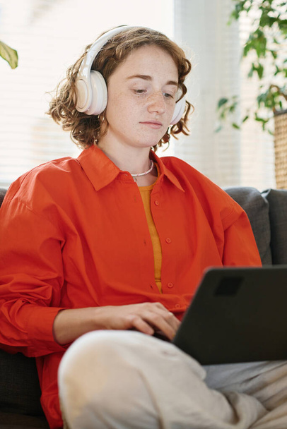 Ciddi üniversite öğrencisi ya da dizüstü bilgisayarı bacaklarında ve kulaklıkları evdeki kanepede oturan serbest çalışan bir kadın. - Fotoğraf, Görsel
