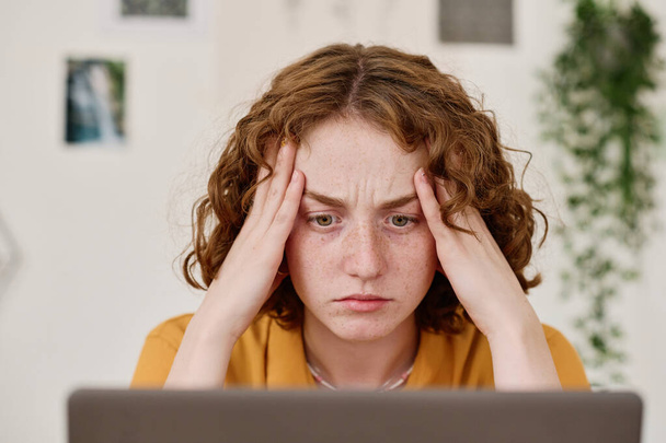 Young female freelancer with tense facial expression looking at laptop screen while sitting by workplace and analyzing information - Photo, Image