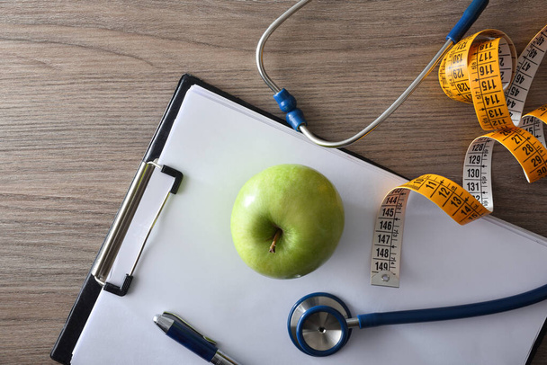 Health control and healthy lifestyle with apple, tape measure and stethoscope on folder with sheets on wooden bench. Top view. - Foto, Imagen