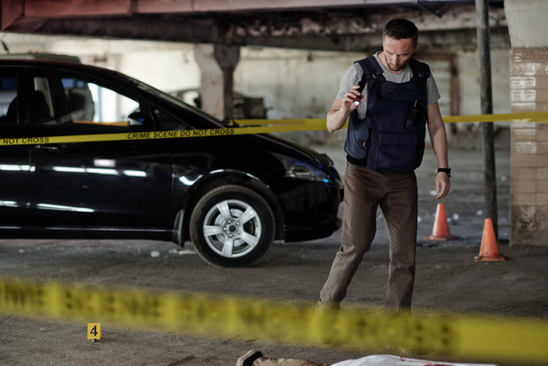 Fiatal férfi bűnügyi helyszínelő keresi a gyilkosság bizonyítékait, miközben egy aszfalton fekvő holttest mellett áll a parkolóban. - Fotó, kép