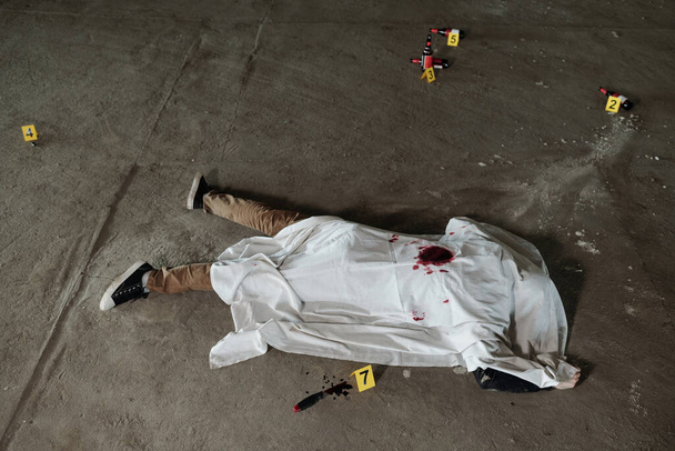 Nad záběrem mrtvého muže pokrytého bílým prostěradlem ležícím na asfaltu parkoviště nebo na nějakém jiném venkovním místě - Fotografie, Obrázek