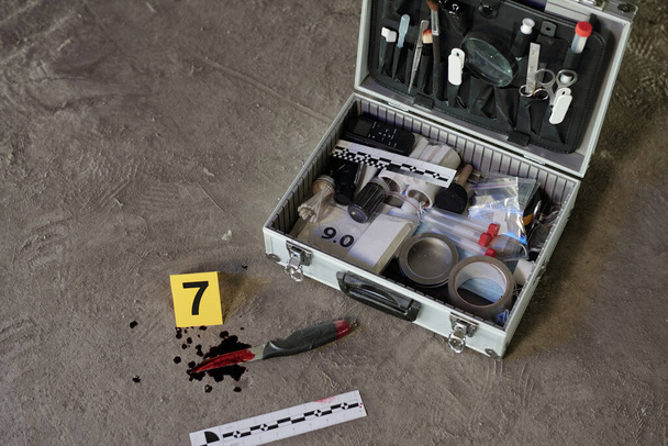 Φονικό όπλο με αίμα θύματος και ανοιχτή βαλίτσα με ποικιλία προμηθειών του ερευνητή της σκηνής του εγκλήματος στην άσφαλτο του χώρου στάθμευσης - Φωτογραφία, εικόνα