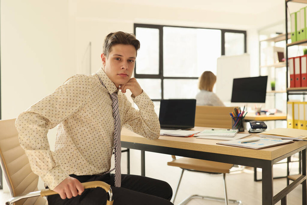 Successful manager sits at table near modern laptop in light office looking in camera. Young man in formal outfit poses at workplace - Fotografie, Obrázek