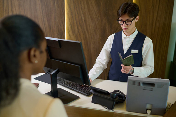 Jonge Aziatische man in uniform en bril controleren van persoonlijke gegevens in paspoort van gasten die voor hem staan in de lounge van het hotel - Foto, afbeelding