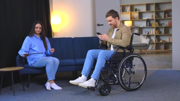 Jovem em cadeira de rodas segurando celular em mãos e sua esposa usando laptop em casa - Filmagem, Vídeo