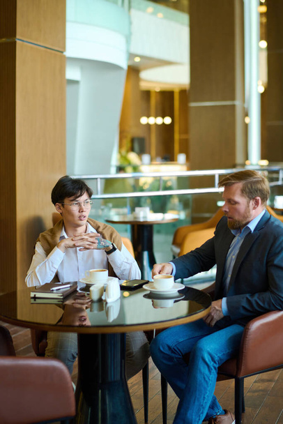 Dois jovens empresários inter-raciais em formalwear tomar café no café ou lounge do hotel moderno e discutir pontos de trabalho - Foto, Imagem