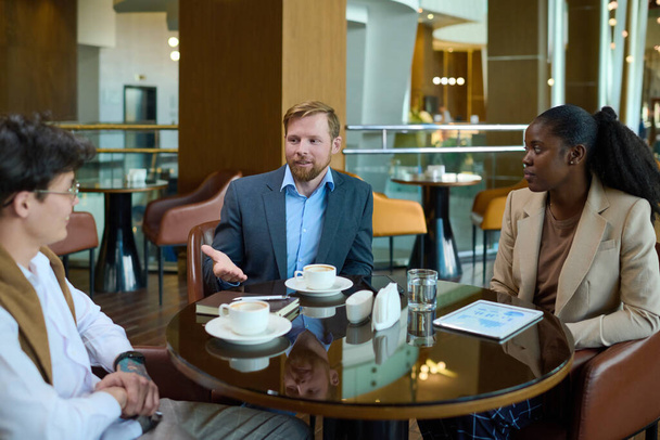 Two young businessmen and black woman in formalwear discussing points of new project while sitting by table in hotel lounge - Photo, Image