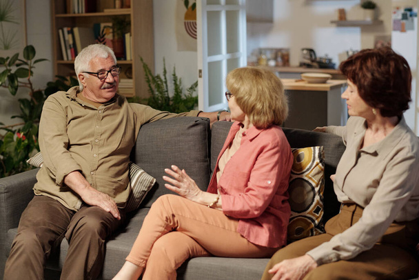 Group of aged friendly people in casualwear discussing latest news while sitting on soft couch in living room during gathering - Photo, image