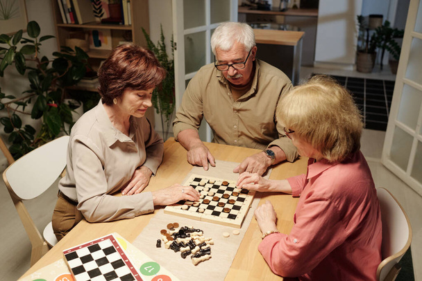 Τρεις συνταξιούχοι φίλοι σε casualwear συγκεντρώθηκαν από το τραπέζι για να παίξουν σκάκι ή checkeres, ηλικιωμένη γυναίκα σκέφτεται την επόμενη κίνηση - Φωτογραφία, εικόνα