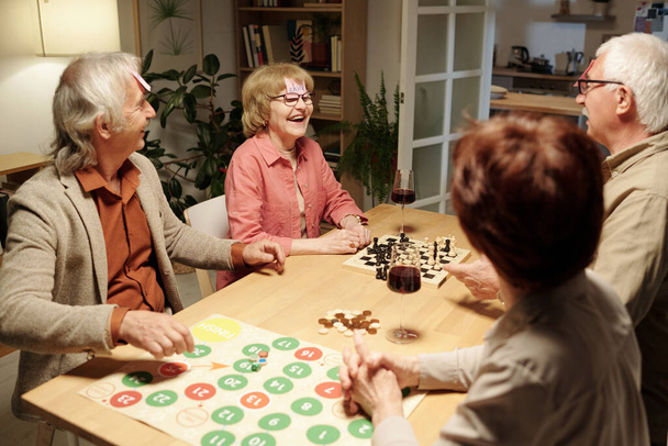 Veselý stárnoucí žena smějící se během názvu hry s přáteli shromážděné u stolu s šachovnicí a papíru se zelenými a červenými kruhy - Fotografie, Obrázek