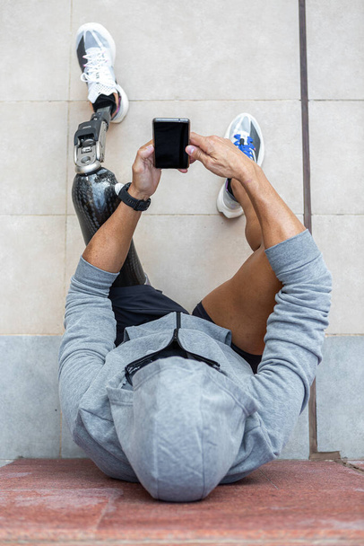 Visão superior de esportista anônimo com perna protética usando celular com tela preta enquanto sentado na calçada perto da parede durante a pausa no treino de fitness durante o dia - Foto, Imagem