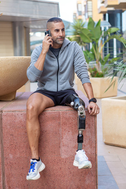 Sportivo di mezza età con gamba protesica che distoglie lo sguardo e risponde alla telefonata mentre è seduto sul blocco di marmo durante la pausa durante l'allenamento di fitness di giorno sulla strada della città - Foto, immagini