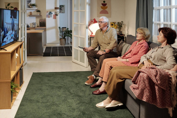 Kolme vanhempaa ystävää katsomassa suosikkisarjaansa istuessaan mukavalla sohvalla television edessä tilavassa olohuoneessa - Valokuva, kuva