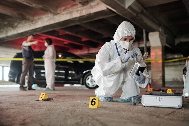 Młody ekspert kryminologiczny w kombinezonach, sprawdzający pustą butelkę na miejscu zbrodni, kucając przed otwartą walizką z materiałami roboczymi. - Zdjęcie, obraz